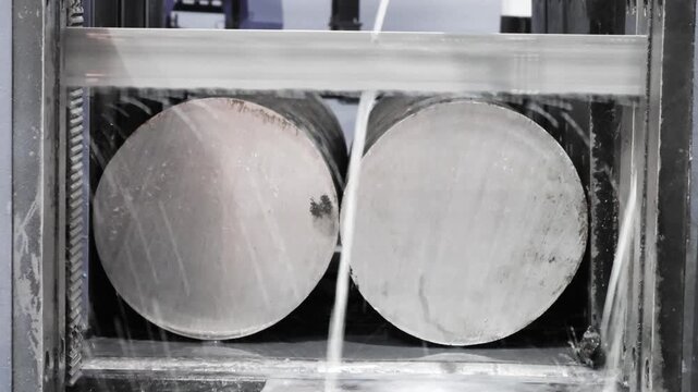 Two metal billets inside a machine enclosure. Water flows around them during processing. Industrial metal shaping process visible through a window
