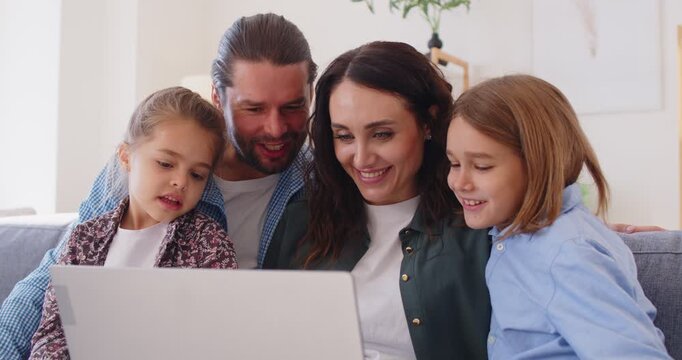 Happy family watching movie online or browsing different websites via laptop computer, sitting on couch and looking at each other happily. Mother, father and two children surfing internet