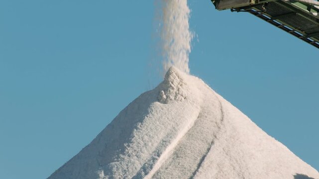 Salt extraction and production with a conveyor belt dumping raw crystals creating a large white mountain under a clear blue sky