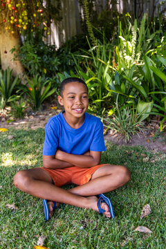 African American child sitting cross-legged on lawn wearing blue T-shirt, orange shorts, flip-flops