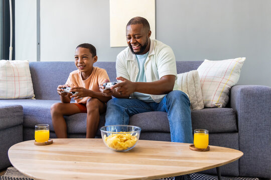 African American father and son sitting on gray sofa playing games with controllers, chips, juice