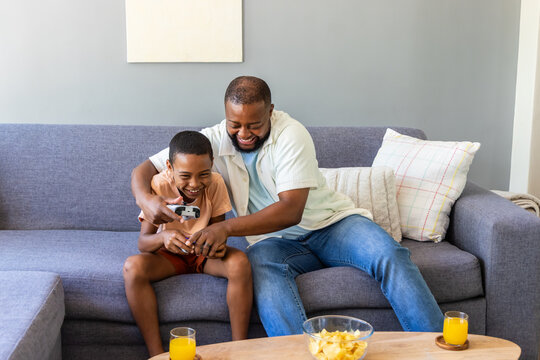 African American dad and teenage son laughing and playing controller on gray sofa with chips, juice