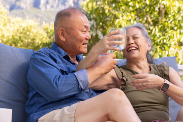 Fototapeta premium Senior Asian couple sitting on outdoor sofa, man holding clear glass, woman in olive top laughing