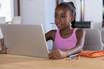 Obraz premium African American child in pink top sitting at dining table, using laptop with orange pencil case