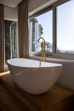 White oval bathtub is sitting on wooden platform in modern bathroom with brass faucet by windows