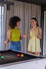 Naklejka premium Two adult women standing chatting at modern lounge by black-felt pool table holding cue ball, cue