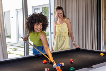 Fototapeta premium Diverse female friends leaning, aiming cue stick on black felt pool table, one wearing sundress