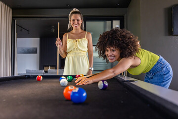 Naklejka premium Diverse female friends playing pool at home with smartphone and cues, lime top, yellow dress