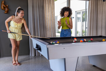 Obraz premium Diverse female friends playing pool on light-gray table, cue stick hitting scattered billiard balls