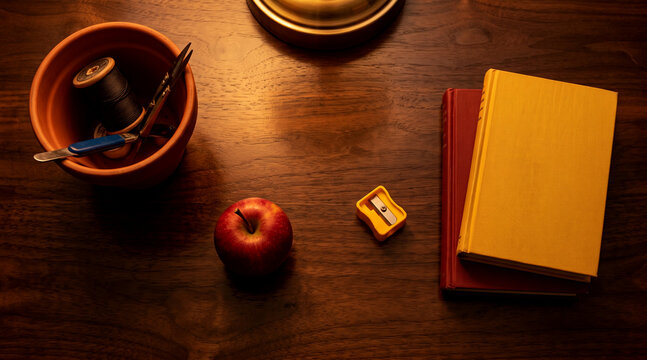 Top down view of a wooden desk featuring books an apple and sewing tools in warm ambient lighting