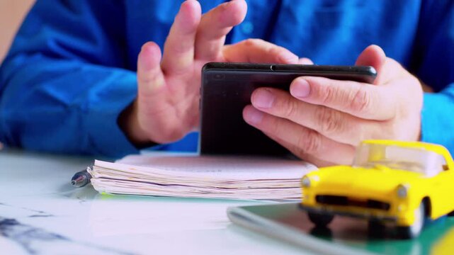 Businessman using car insurance app on digital tablet and writing notes in pad. Focus on hands, yellow toy car on desk. Close up, shallow depth of field, natural daylight. 