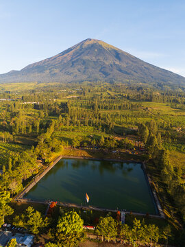 Aerial view of Embung Kledung reservoir with Mount Sindoro in the background, surrounded by lush green agricultural fields and trees in Temanggung Regency, Central Java, Indonesia.