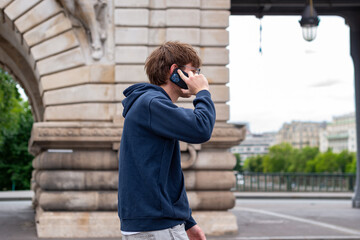 Fototapeta premium A profile view of a young man walking on a city street while talking on a smartphone. He wears a dark hoodie and holds the mobile device to his ear during daytime.