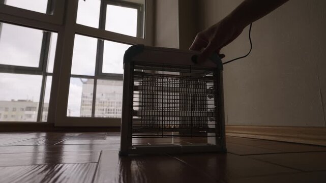 Person hand turning on electric insect zapper with glowing blue ultraviolet lamps standing on wooden floor to control pests and flying mosquitos inside room with large window. Shooting in slow motion.