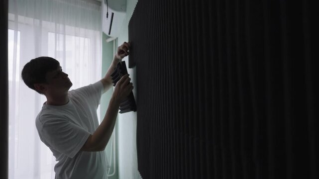 Young man installing black acoustic foam panels on wall to improve room acoustics and sound insulation for home recording studio, reducing noise echo and reverberation. Shooting in slow motion.