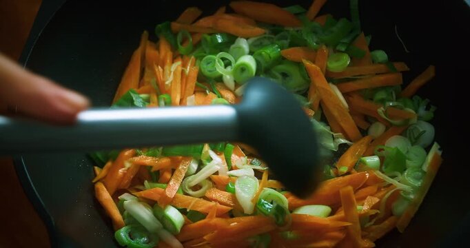 Close up of a chef stirring sliced carrots and leeks in a pan using a silicone spoon. Fresh vegetables being sauteed during homemade meal preparation in the kitchen.