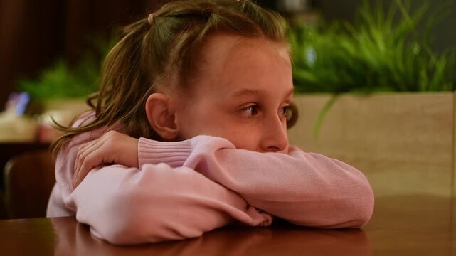 Young girl looks bored with head resting on folded arms at cafe table while waiting for food during family dining visit.