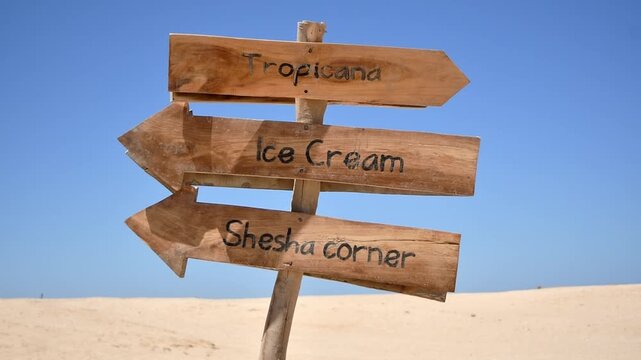 Rustic wooden signs guide tourists toward tropical attractions and refreshments under clear blue sky at sunny beach resort.