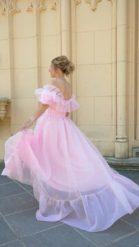 Beautiful happy blonde female in luxury pink ruffles dress twirling and smiling near historical building wall. Vertical video format.