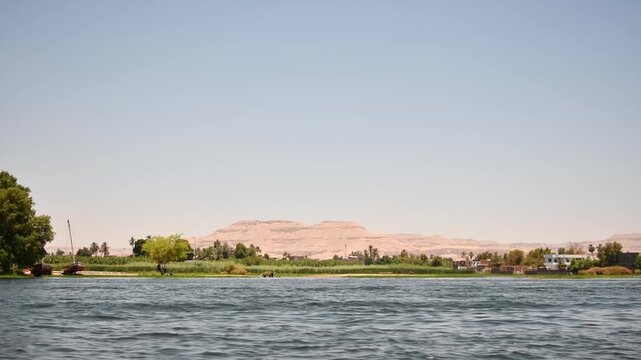 Boat ride along Nile River shows green shore, village houses and desert hills beneath wide Egyptian sky on warm sunny day.