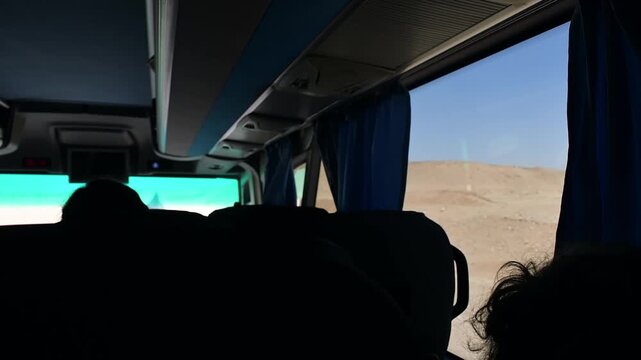 Tourists ride sightseeing bus through arid desert landscape, watching sunlit hills through large windows during excursion.