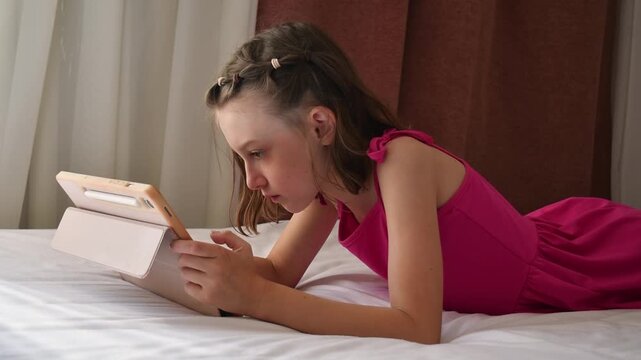Focused girl watches online content on tablet while lying on hotel bed during quiet family vacation and indoor leisure time.