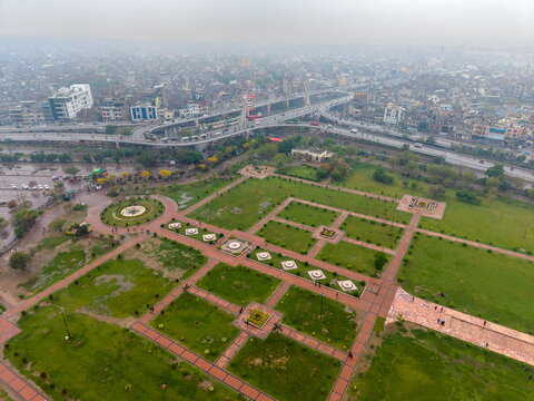 Aerial view of Minar-e-Pakistan and Greater Iqbal Park with green lawns, geometric pathways, and a busy elevated highway overpass in the background in Lahore, Punjab, Pakistan.