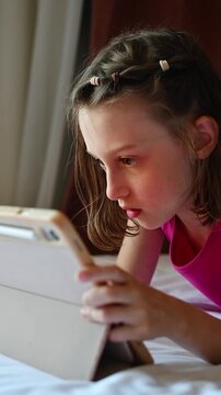 Focused girl plays touchscreen game on tablet in bed during indoor leisure, absorbed in digital entertainment and screen time