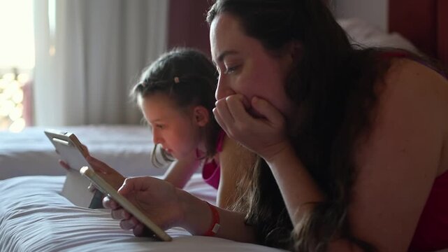 Mother and daughter relax on hotel bed and use smartphone and tablet together during quiet family leisure time on vacation.