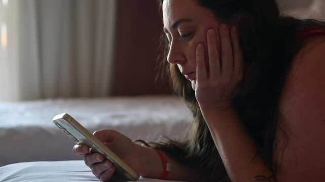 Young woman lies on hotel bed and scrolls smartphone with bored expression during quiet downtime alone in softly lit room.