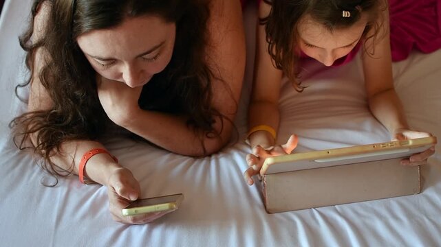 Mother and daughter use smartphone and tablet on hotel bed during shared family leisure time while traveling abroad together.
