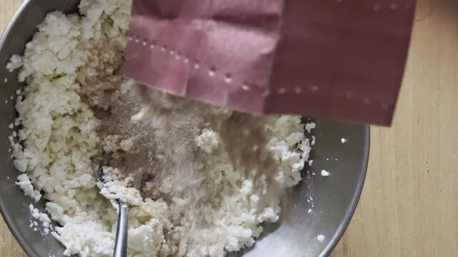 Overhead shot of pouring brown powder ingredient from paper packaging into cheese mixture