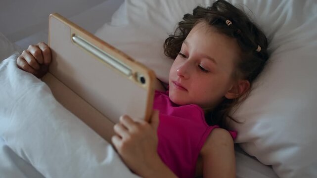 Young girl uses tablet under white duvet in bed and enjoys calm leisure time with digital entertainment during indoor rest.