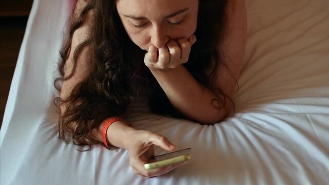 Young woman lies on hotel bed, rests chin on hand and looks at smartphone while relaxing alone during quiet leisure time.