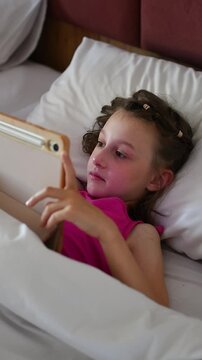 Young girl watches cartoons on tablet while lying under white duvet in bed during quiet indoor leisure and screen time.