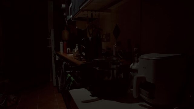 A dimly lit kitchen inside a small New Zealand studio apartment during the early morning. Soft natural light begins to enter the room while countertops, cookware