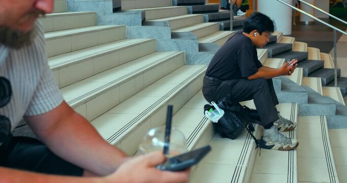 Man sitting on stairs using smartphone while another person holds phone and drink in foreground, indoor staircase setting with diagonal steps, casual waiting, visible hand gestures and mobile use