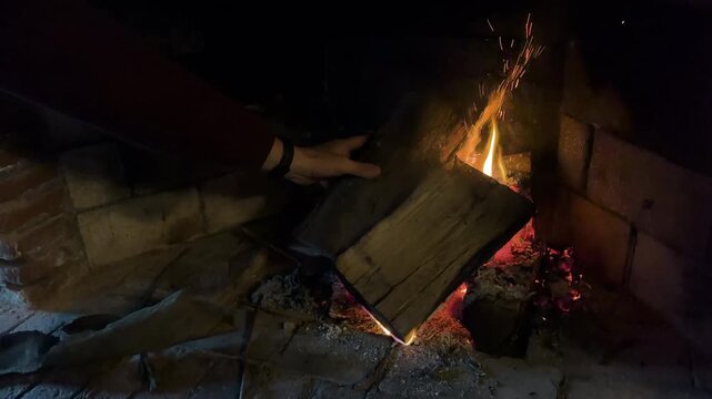 Close up of a person adding a wooden log into a warm burning fireplace to stoke the fire on a cold winter day
