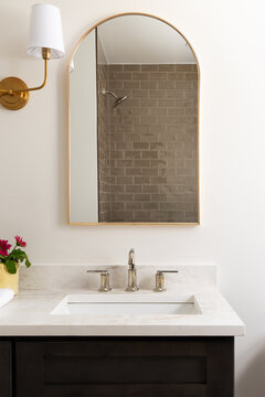 A bathroom detail with a marble countertop on a dark wood vanity cabinet. gold faucet and light fixture, and a brown subway tiled shower.
