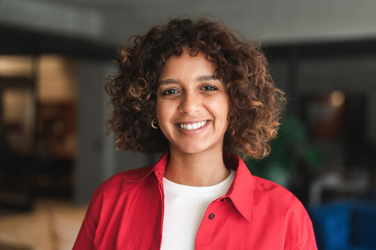 Beautiful latin or muslim arabian young woman businesswoman with curly hair looking at camera indoors in office, lifestyle portrait. Indian girl teenager student smiling standing in university campus