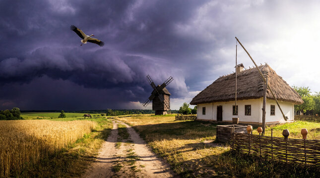 Traditional Ukrainian authentic cottage and wooden windmill under dramatic storm clouds with flying stork