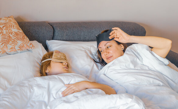 Close up eye level shot of a mother sleeping with a black eye mask and her preteen daughter hiding under white sheets with a beige fuzzy sleep mask on a bed