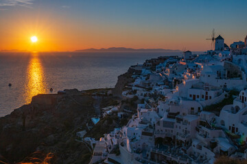 Golden sun reflecting on Aegean Sea beside whitewashed hillside homes and windmill in Oia at dusk. © Andrei Tomas