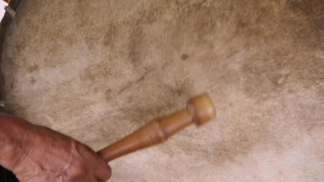 Hand strikes drum with wooden mallet. Drum skin shows natural, aged texture. Beat pulses rhythmically with steady hand. Cultural music practice captured in motion. Evokes tradition, earthy. Lamas, Tar