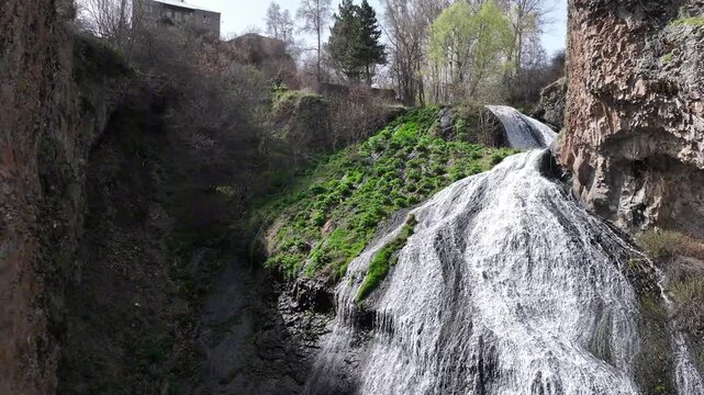 An aerial drone shot captures the majestic Jermuk Waterfall, known as Mermaid's Hair, cascading down rocky cliffs amidst lush green vegetation in Armenia's beautiful mountain landscape during spring.
