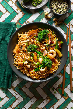 Tasty teriyaki chicken bowl with egg noodles, broccoli, zucchini, bell pepper, and sesame seeds in a black bowl on a green and white tiled background.