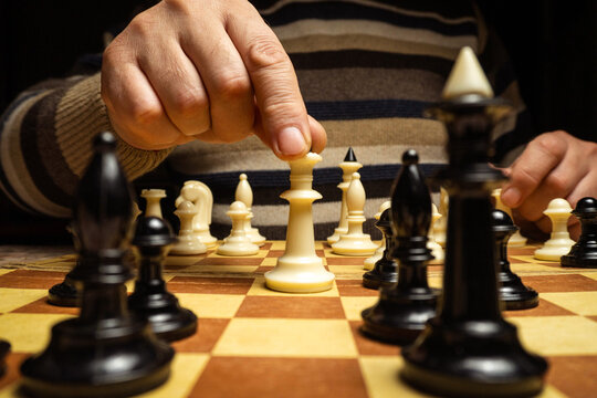 A person focuses on a chess match and moves a white piece on a wooden chessboard. Black and white pieces fill the board as the game unfolds