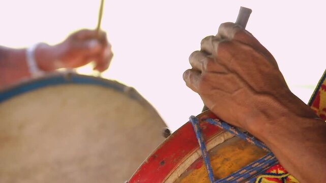 Hands beat drum with rhythm and passion. Drum skin taut, painted with bold colors. Cultural celebration pulses through steady beats. Sunlight glows softly behind musicians. Lamas, Tarapoto. Peru.