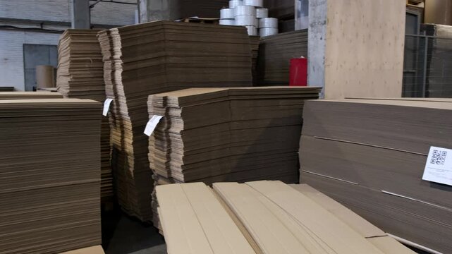 Large volume of brown corrugated cardboard sheets neatly stacked on wooden pallets prepared for box production in a factory workshop.