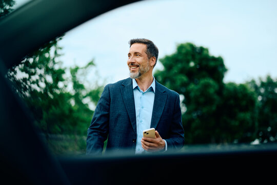 Happy businessman using cell phone standing near his car outdoors.